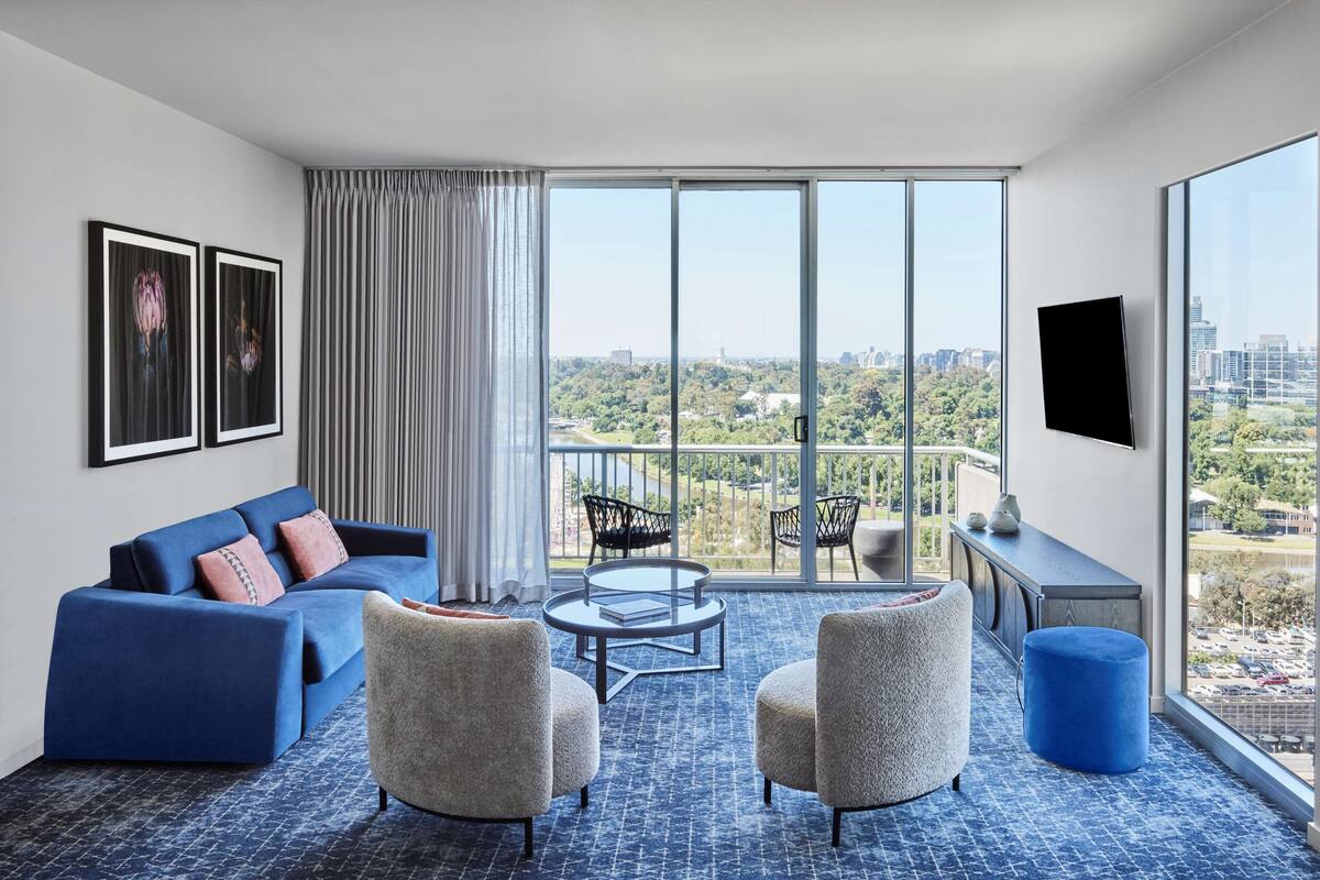 A modern hotel room with a large window, a sofa, two armchairs, a small round table, and a TV on the wall.