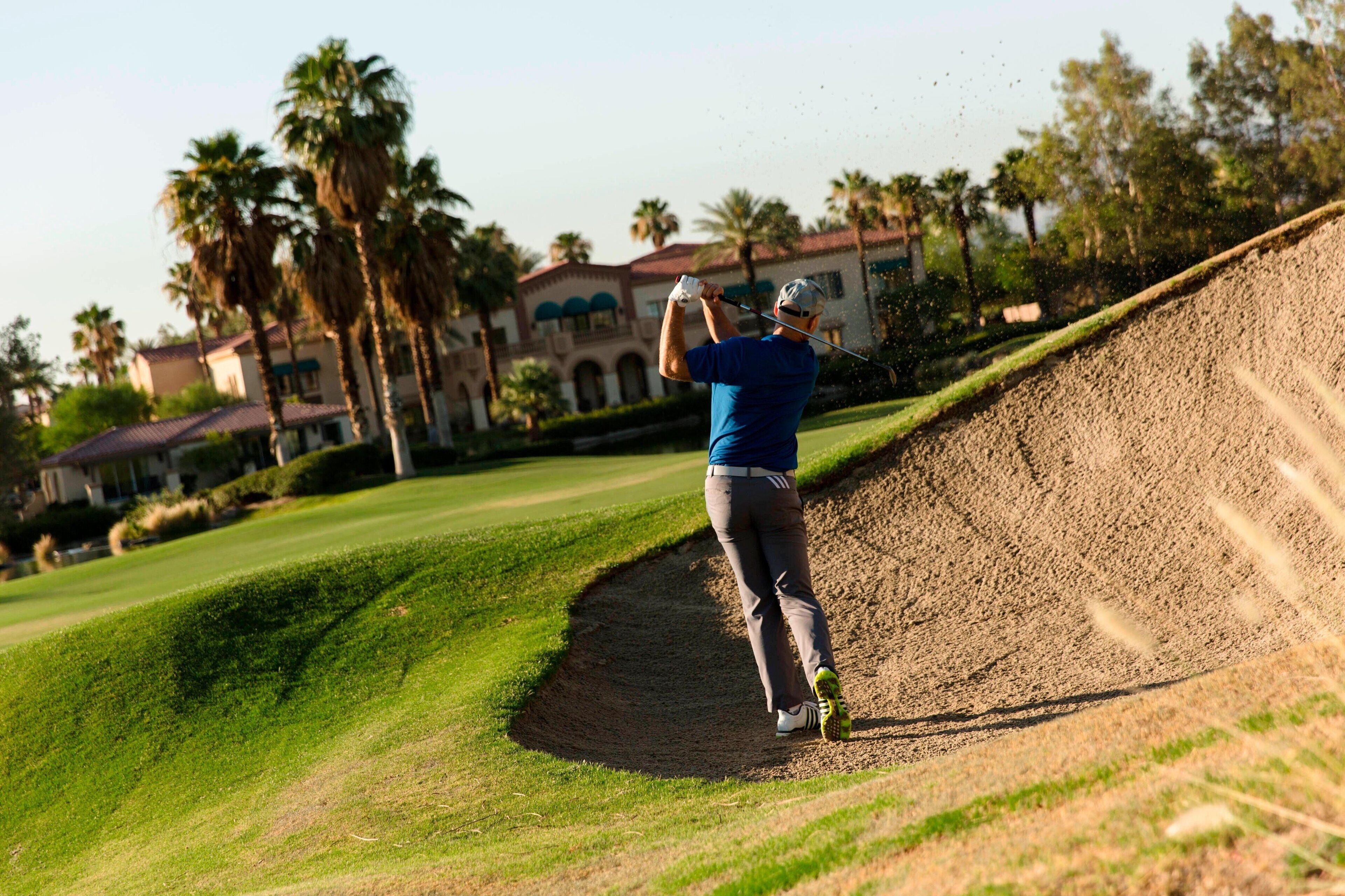 Un golfista en un campo de golf con un búnker de arena, palmeras y un edificio al fondo.