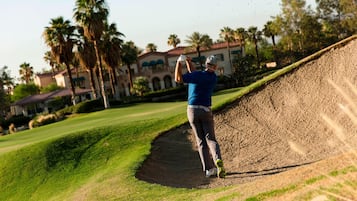 Un golfista en un campo de golf con un búnker de arena, palmeras y un edificio al fondo.