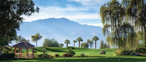 A golf course with a gazebo, palm trees, and mountains in the background.