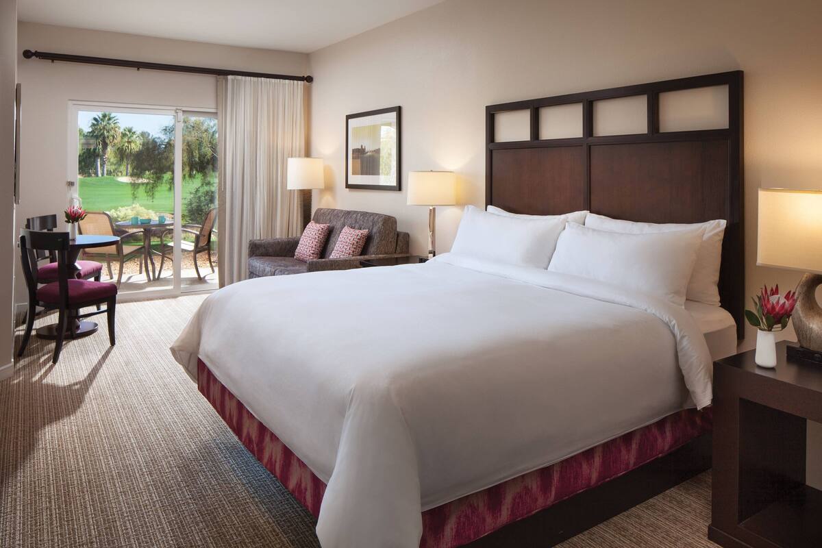 A hotel room with a large bed, a desk with a chair, a sofa, and a view of a golf course through the window.