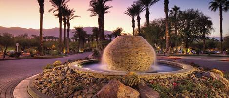 A circular fountain with a stone structure in the center, surrounded by a landscaped area with rocks and cacti, palm trees, and a mountainous backdrop.