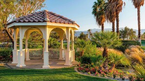 A gazebo with a tiled roof and white columns, surrounded by a well-maintained garden with palm trees and flowering plants.