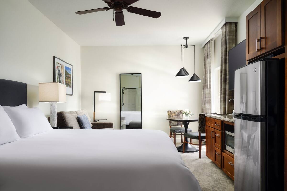 Habitación de hotel con una cama grande, ventilador de techo, kitchenette con refrigerador y comedor con mesa y sillas.