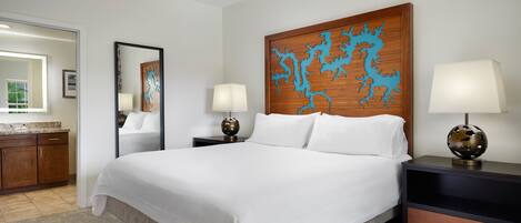 A hotel room with a large bed, two bedside tables with lamps, a wooden headboard with a blue design, a ceiling fan, and a bathroom visible through an open door.