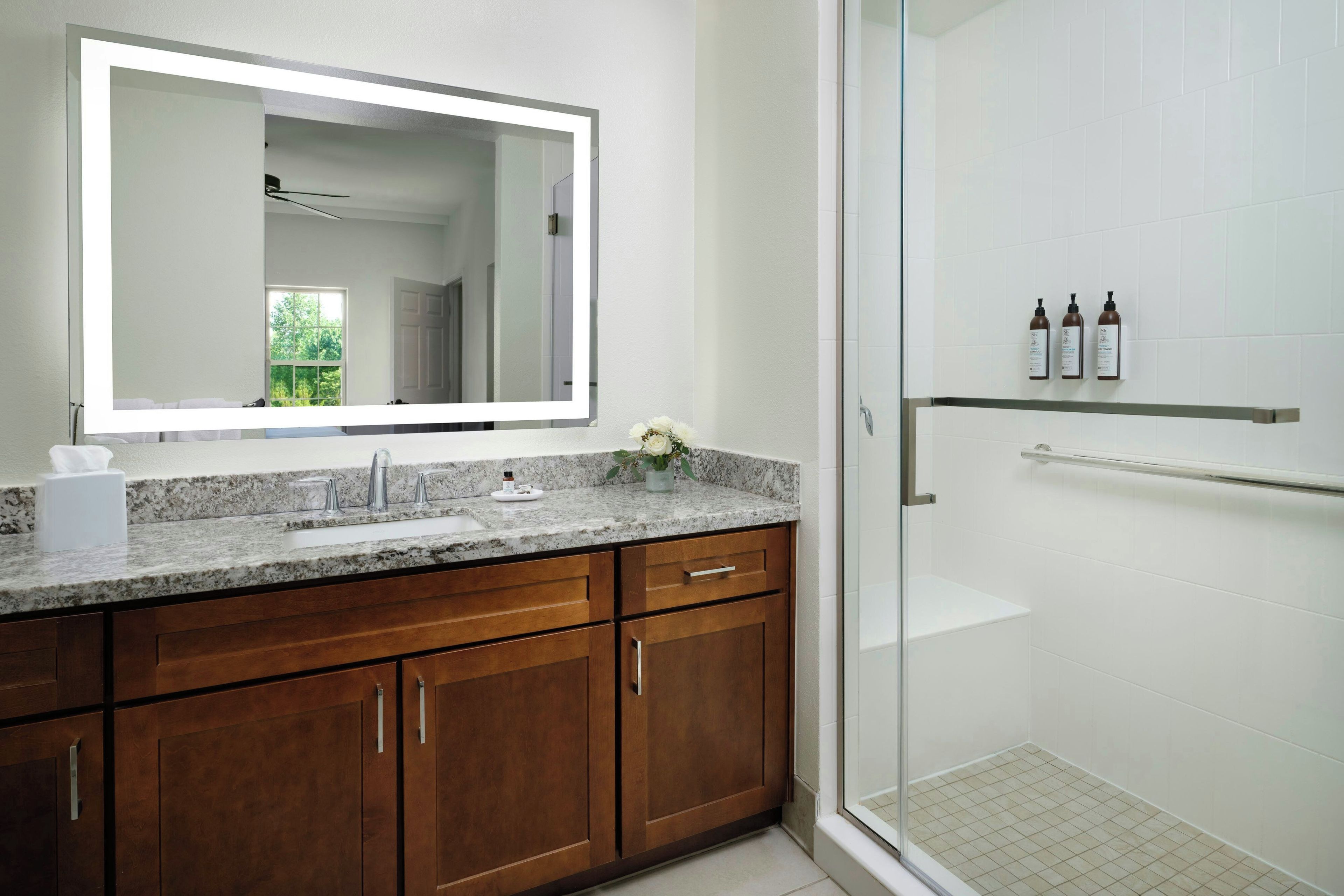 A modern bathroom with a large mirror, granite countertop, wooden cabinets, and a glass shower enclosure.