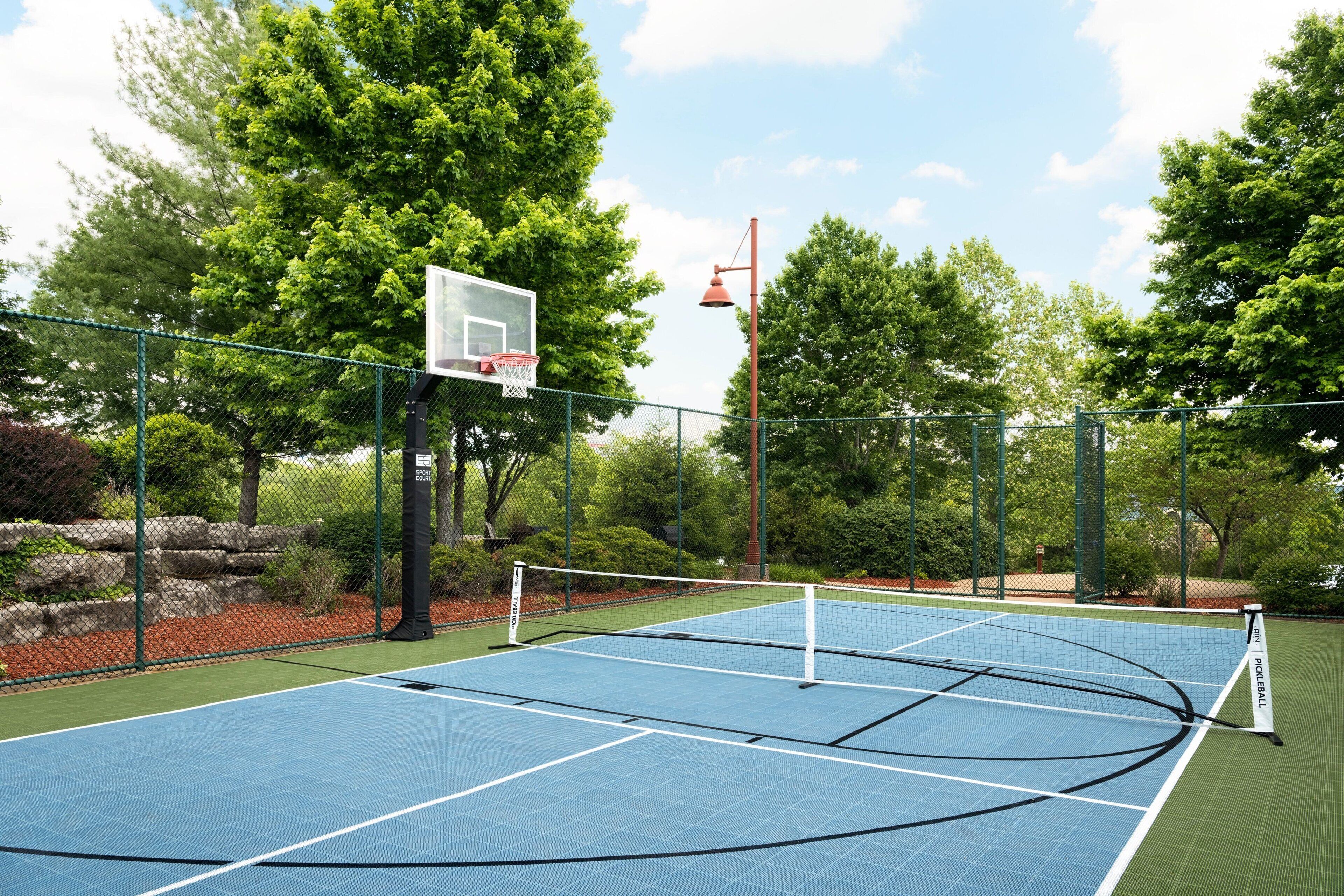 An outdoor tennis court with a net, surrounded by trees and a basketball hoop.