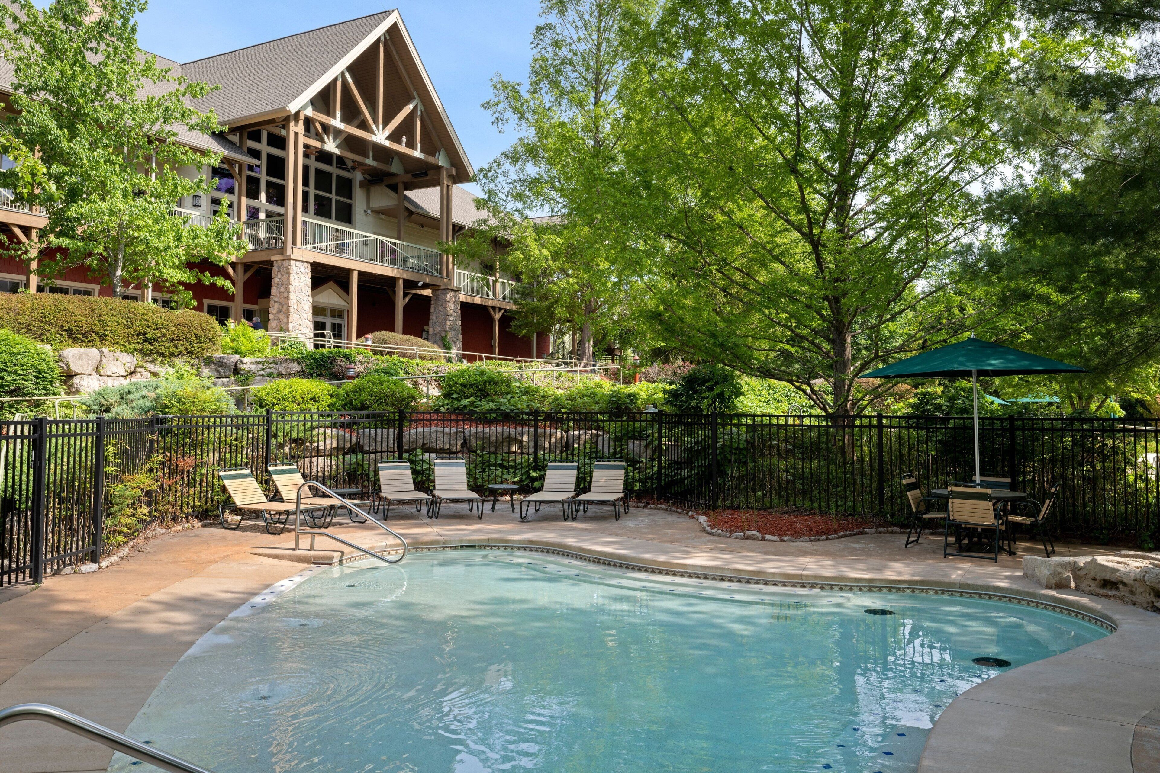 A swimming pool with lounge chairs and a covered patio area, surrounded by a fence and trees.