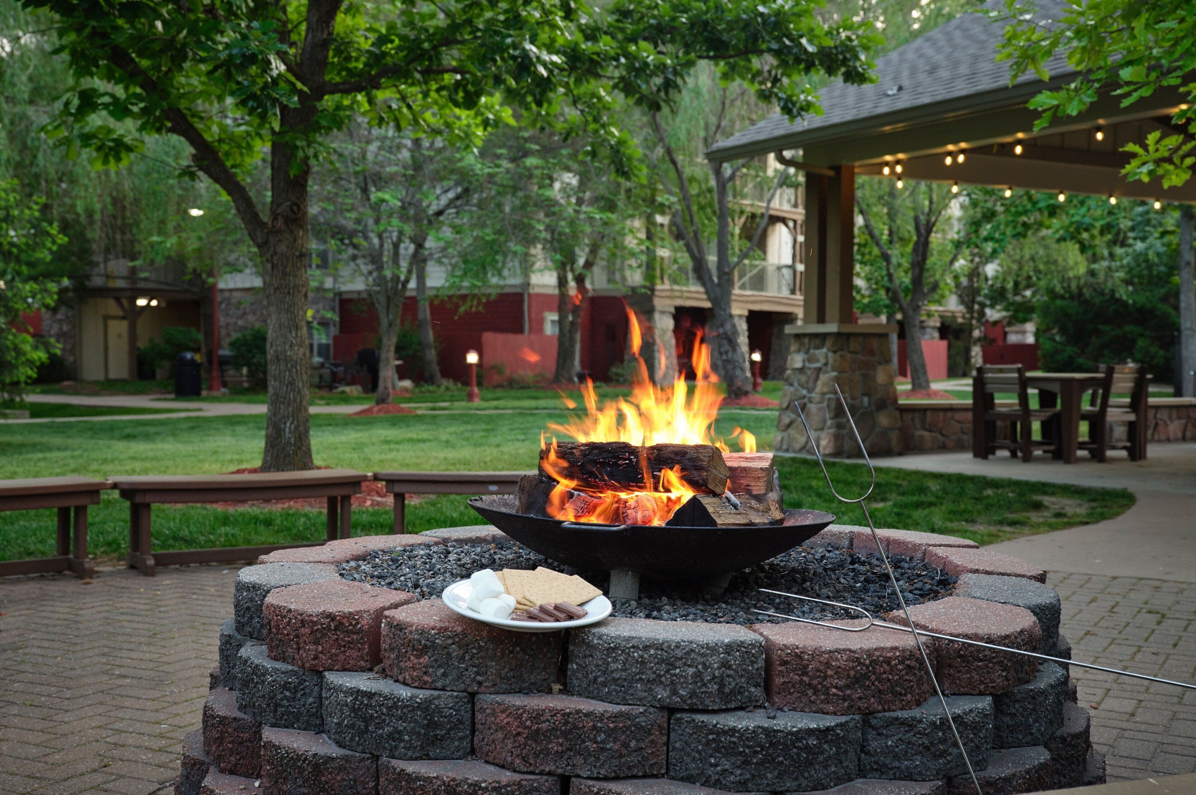 A fire pit with a plate of marshmallows and s'mores, surrounded by stone blocks, in a landscaped outdoor area with benches and a covered seating area in the background.