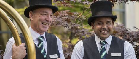 Two men in formal attire with name tags, standing outdoors with a blurred background of greenery.