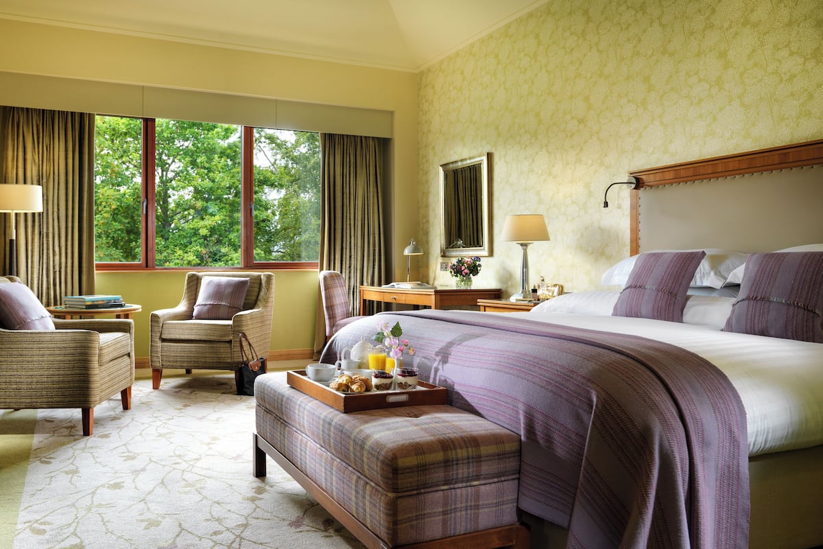 A hotel room with a large bed, a sitting area with a chair and a small table, a dresser, and a window with curtains.