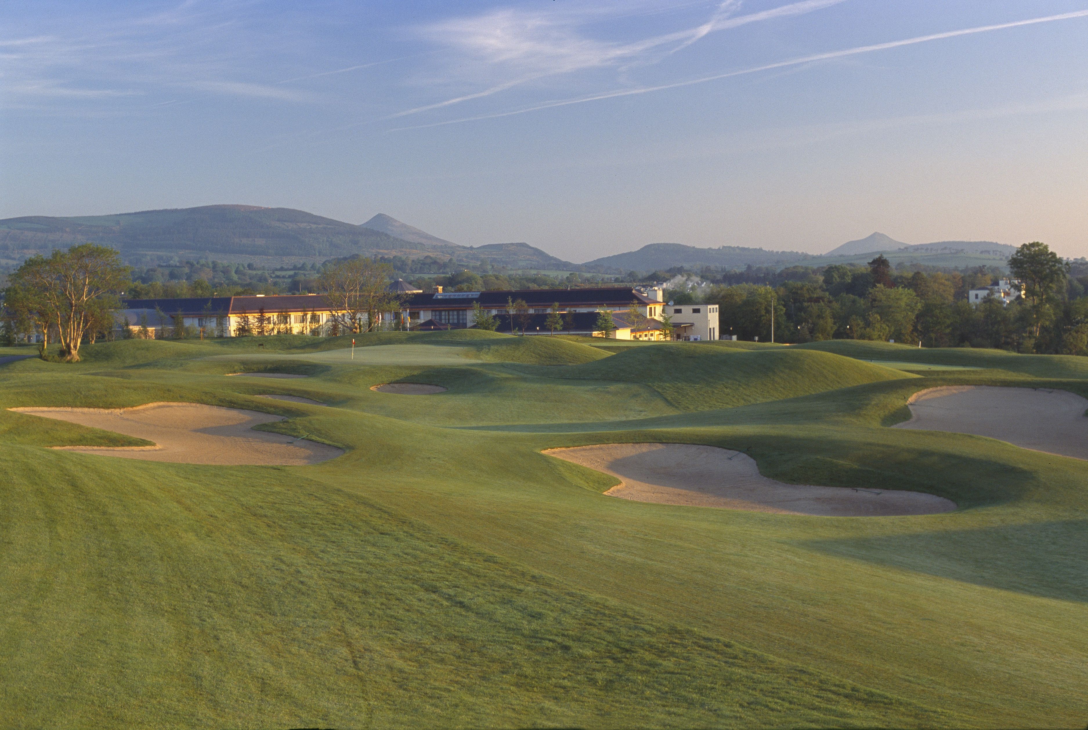 A golf course with bunkers, a clubhouse, and distant mountains.