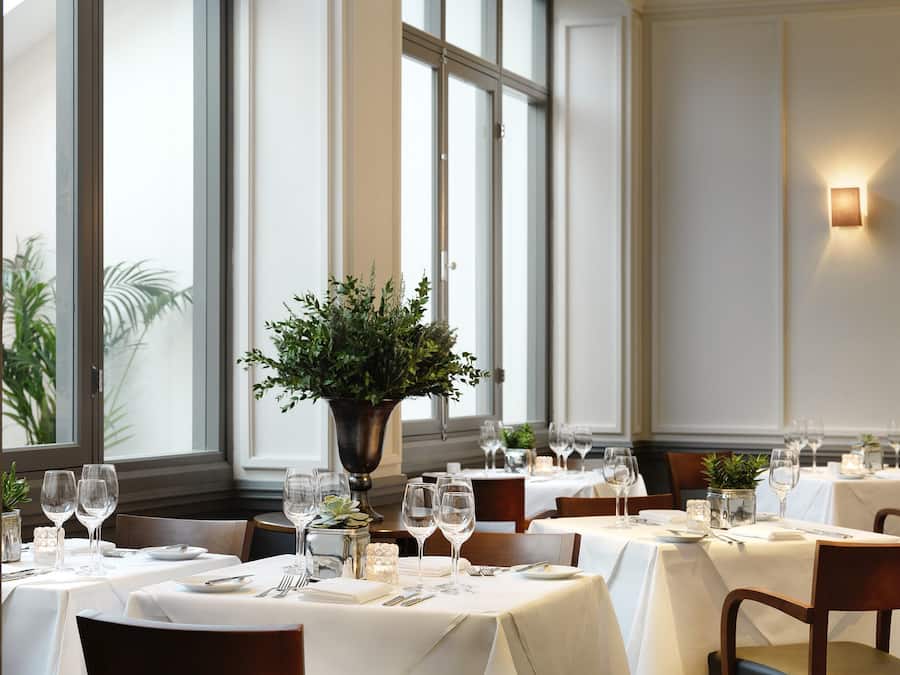 Un restaurante con varias mesas dispuestas para comer, con manteles blancos, copas de vino y un centro de mesa con planta en maceta.
