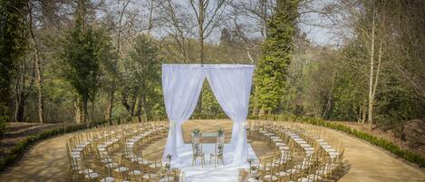 Montaje para una ceremonia de boda al aire libre con un dosel blanco, mesas redondas y sillas dispuestas en semicírculo.