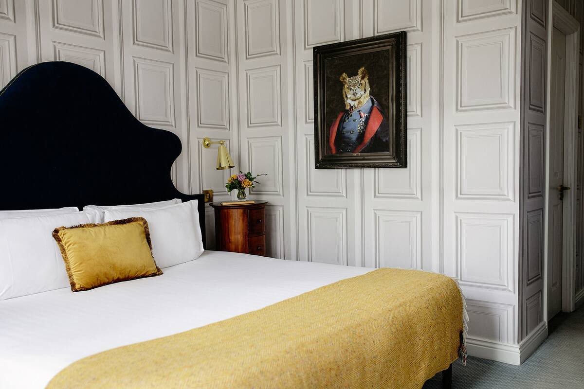 A neatly made bed with a black headboard, a gold pillow, and a yellow throw blanket. There is a framed picture on the wall and a small wooden nightstand with a lamp and a vase of flowers.