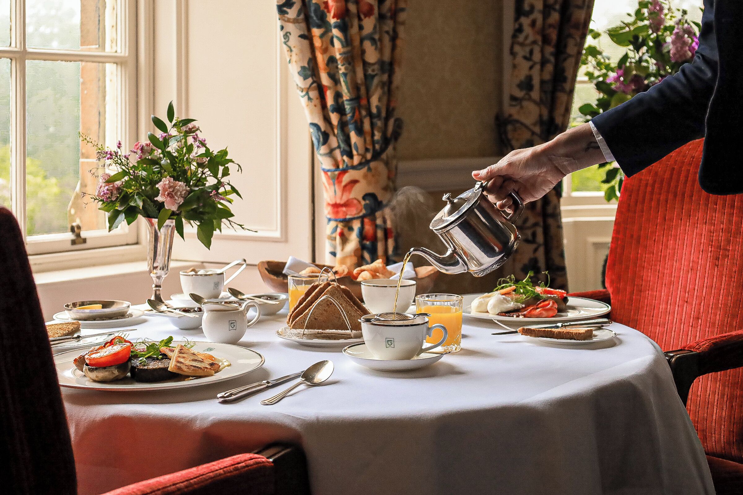 Une table de petit-déjeuner dressée avec des assiettes remplies de nourriture, une théière versant du thé et un vase de fleurs.