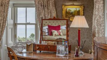 A room with a wooden table, a chair, a bed with a red pillow, a lamp, and a framed painting on the wall.