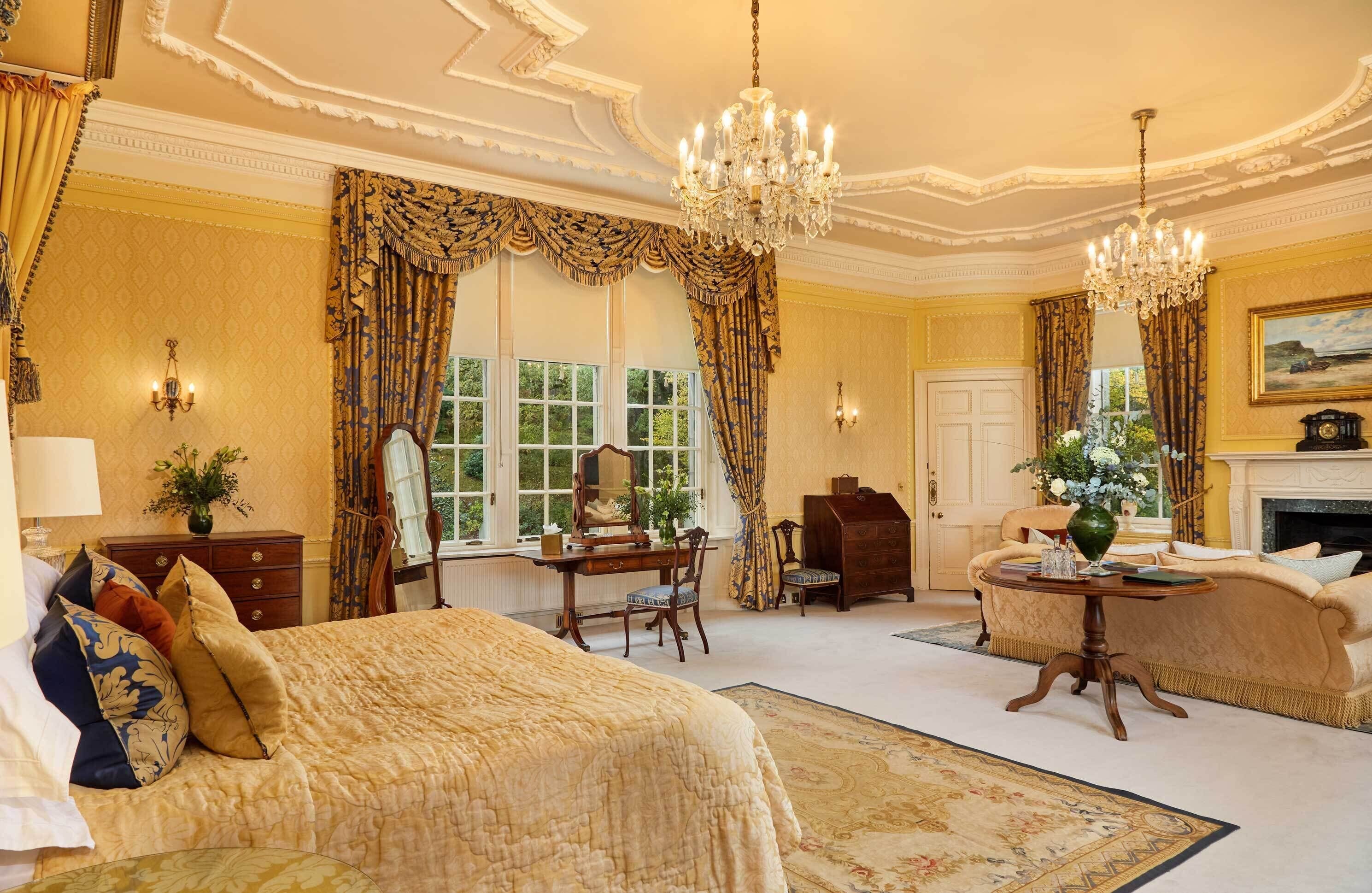 Une chambre à coucher luxueuse, dotée d’un grand lit, d’un bureau, d’une chaise et d’un canapé. On y trouve un lustre, une cheminée et une grande fenêtre avec des rideaux.