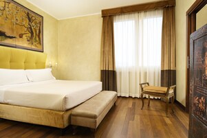 A hotel room with a large bed, a bench, a wooden chair, a painting on the wall, and a window with curtains.