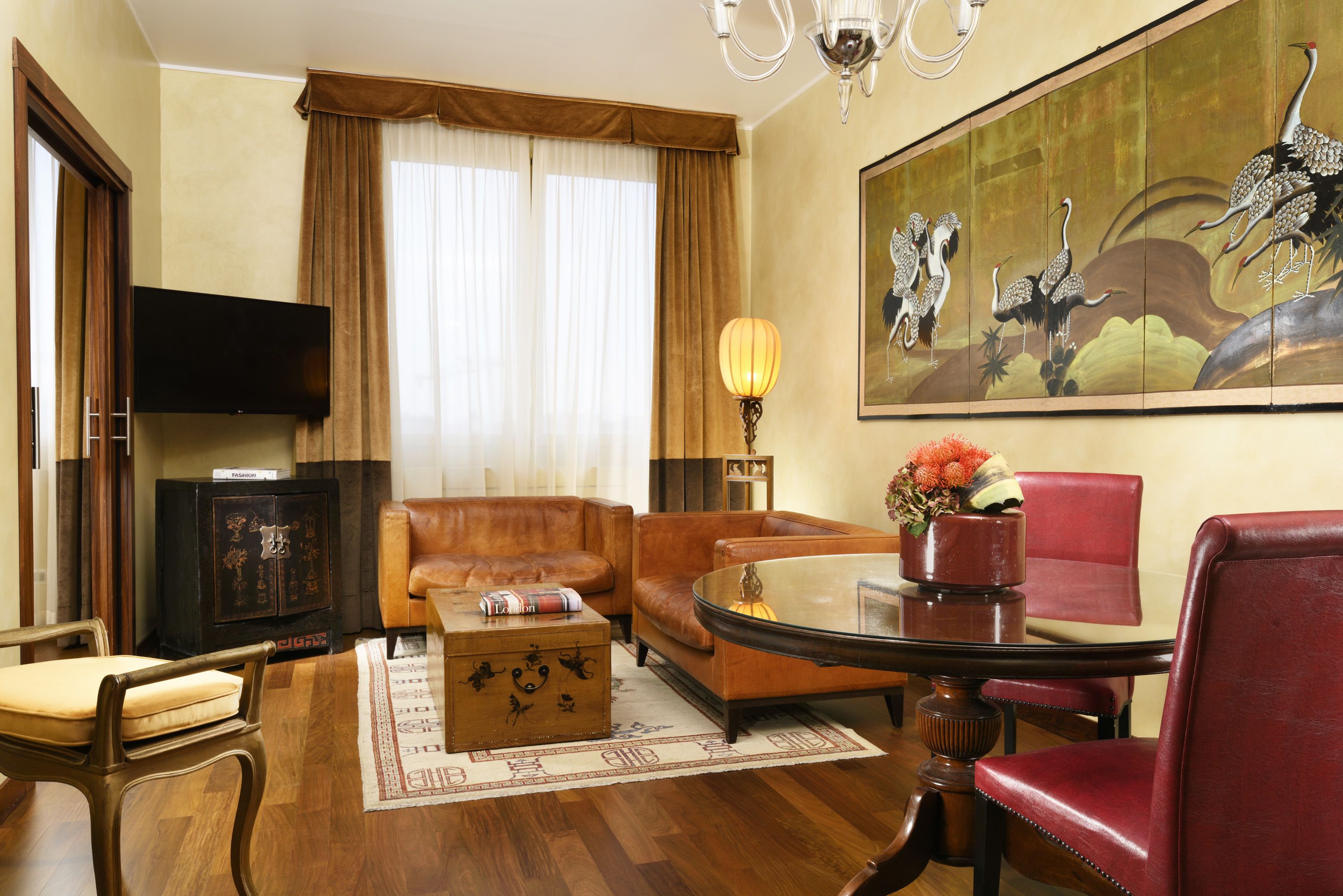 Een hotelkamer met een bank, fauteuil en een eettafel met stoelen. Er is een televisie aan de muur bevestigd en een schilderij boven de bank.