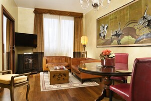 Een hotelkamer met een bank, fauteuil en een eettafel met stoelen. Er is een televisie aan de muur bevestigd en een schilderij boven de bank.
