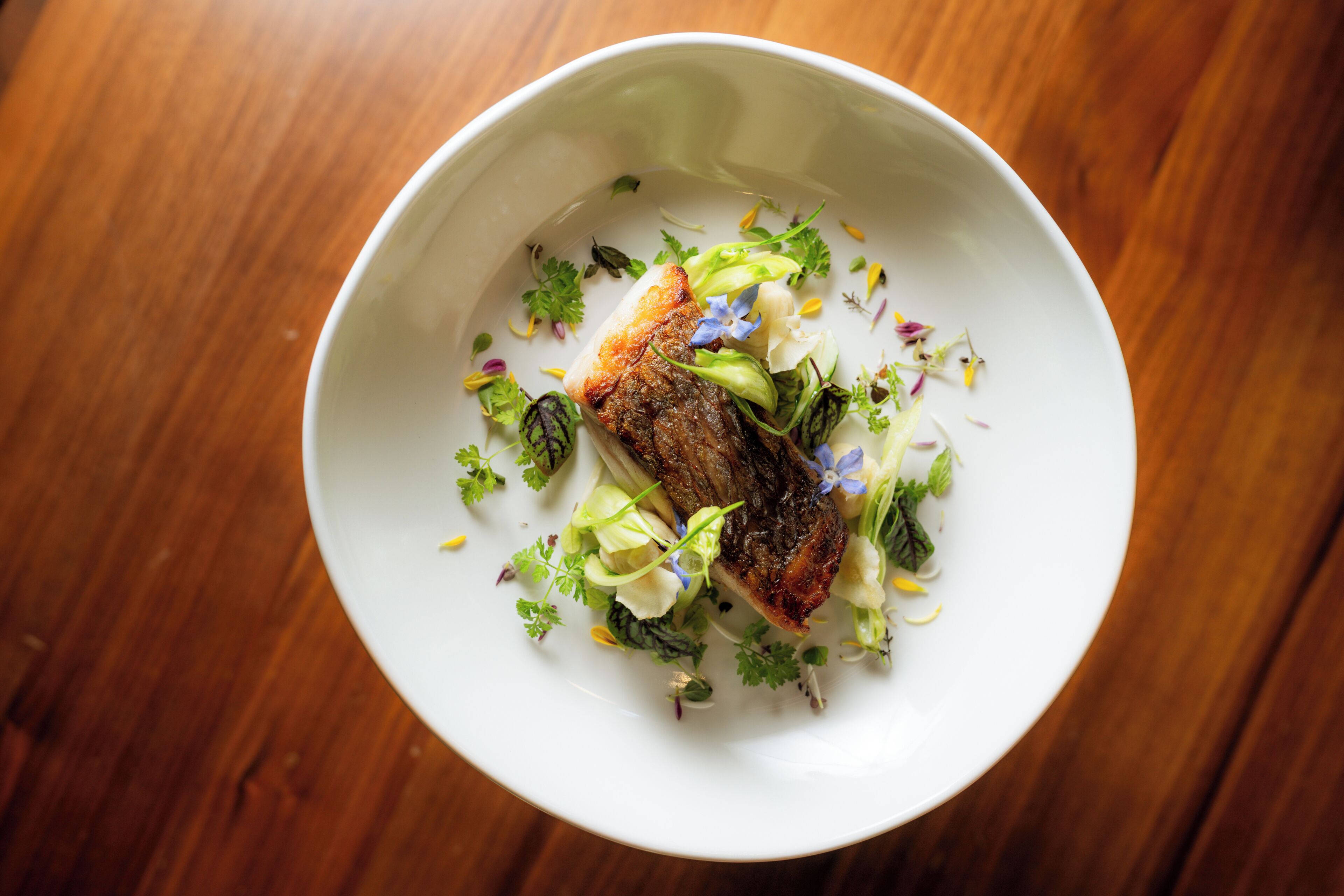 A white bowl with a piece of meat and greens on a wooden table.