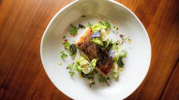 A white bowl with a piece of meat and greens on a wooden table.