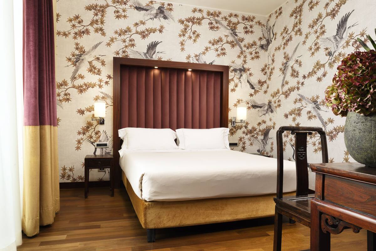 A hotel room with a bed, wooden floor, a nightstand, a lamp, and a decorative vase.