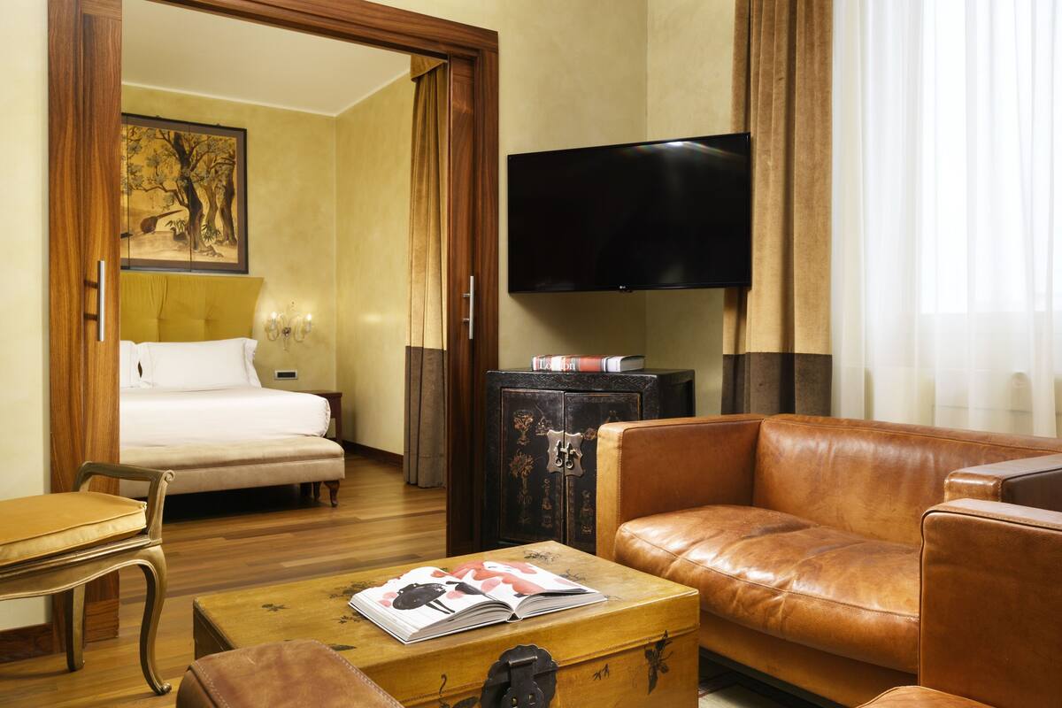 A hotel room with a bed, a leather sofa, a television, and a wooden chest of drawers.