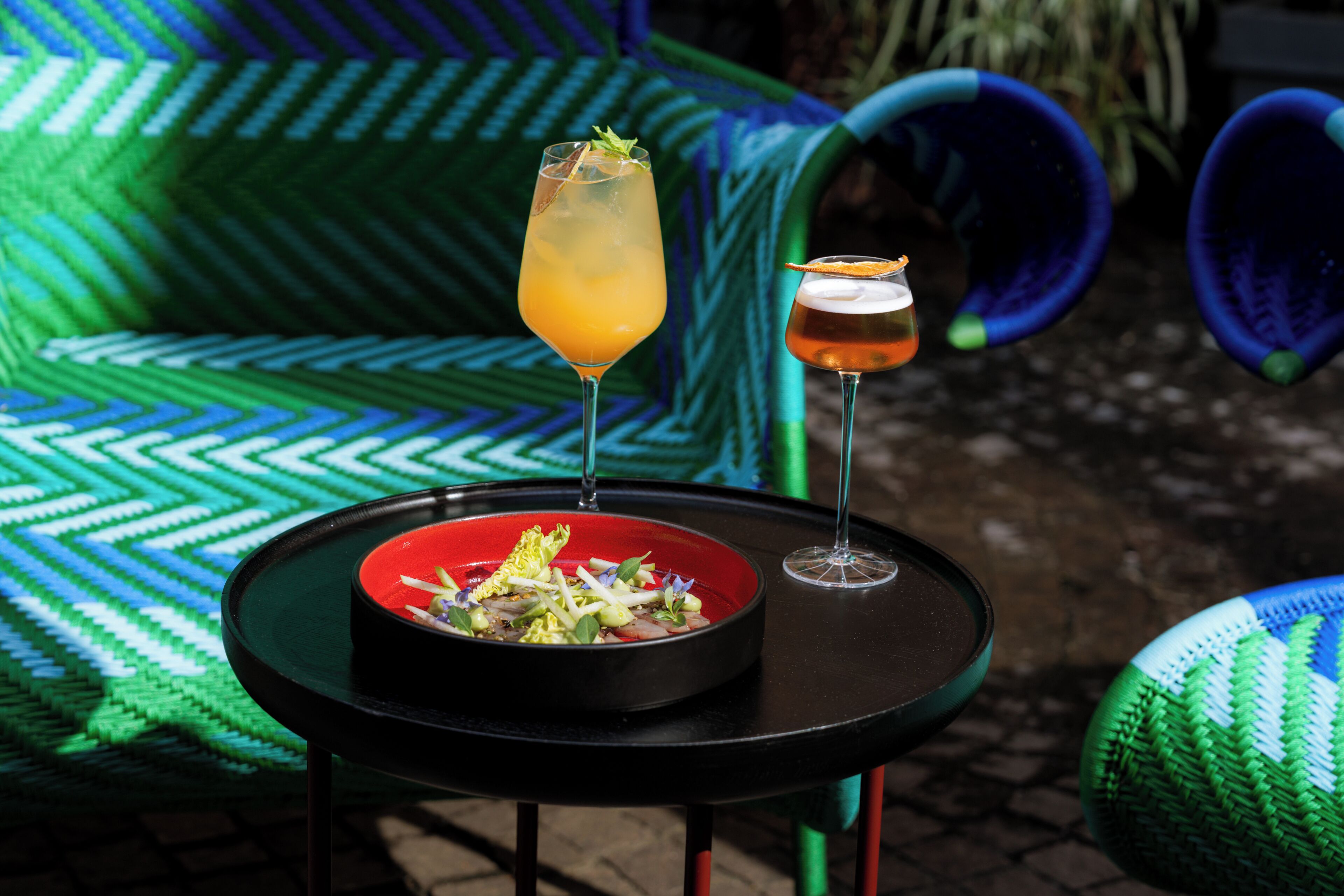 A table with a salad bowl and two drinks, a chair with a woven pattern, and a woven floor mat.
