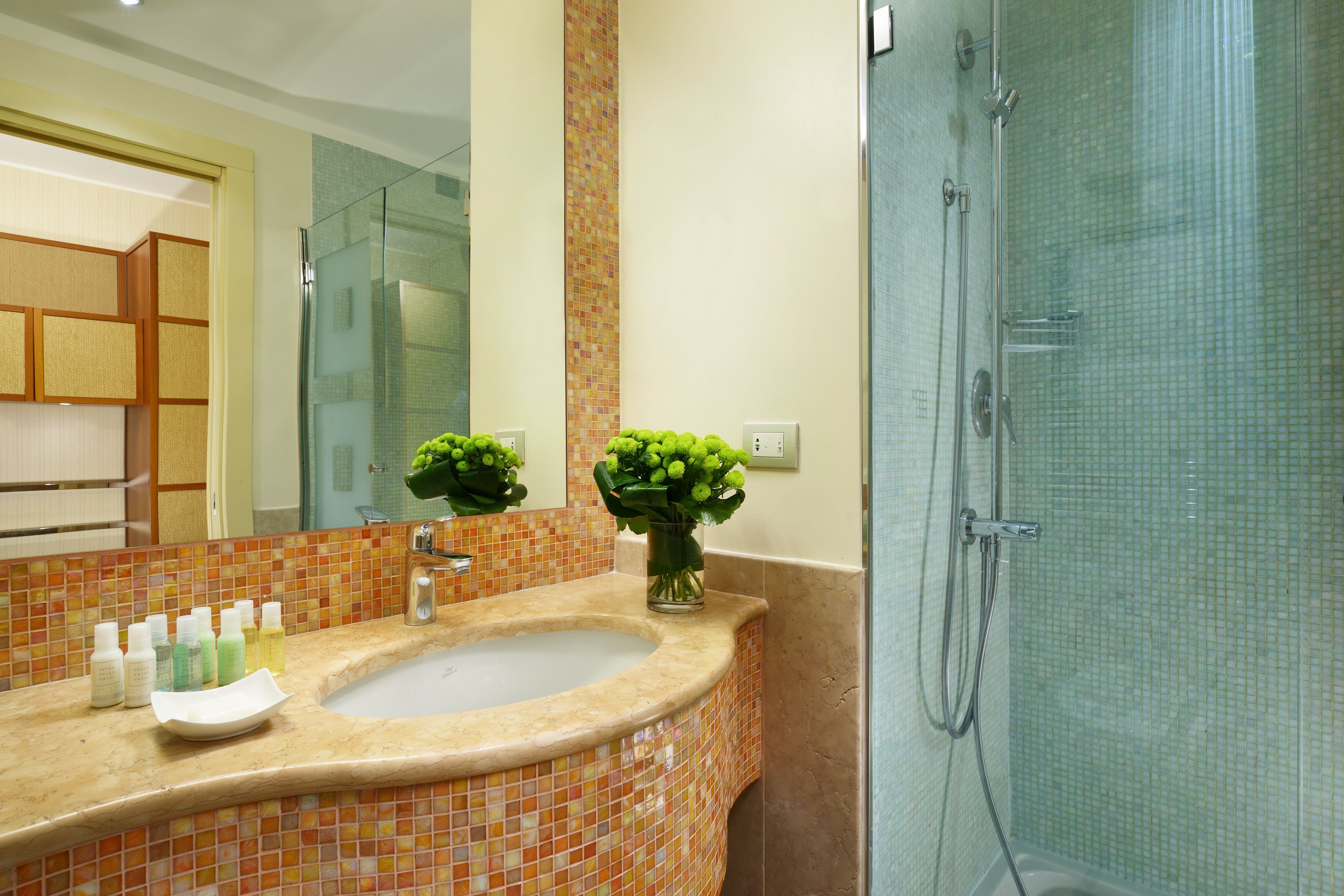 Ein Badezimmer mit Waschbecken, Spiegel, Dusche und einer Vase mit Grünpflanzen.