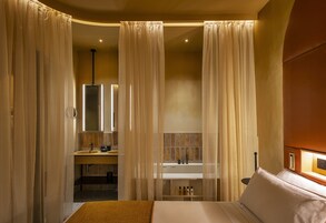A hotel room with a bed, a bathroom with a sink and mirror, and sheer curtains.