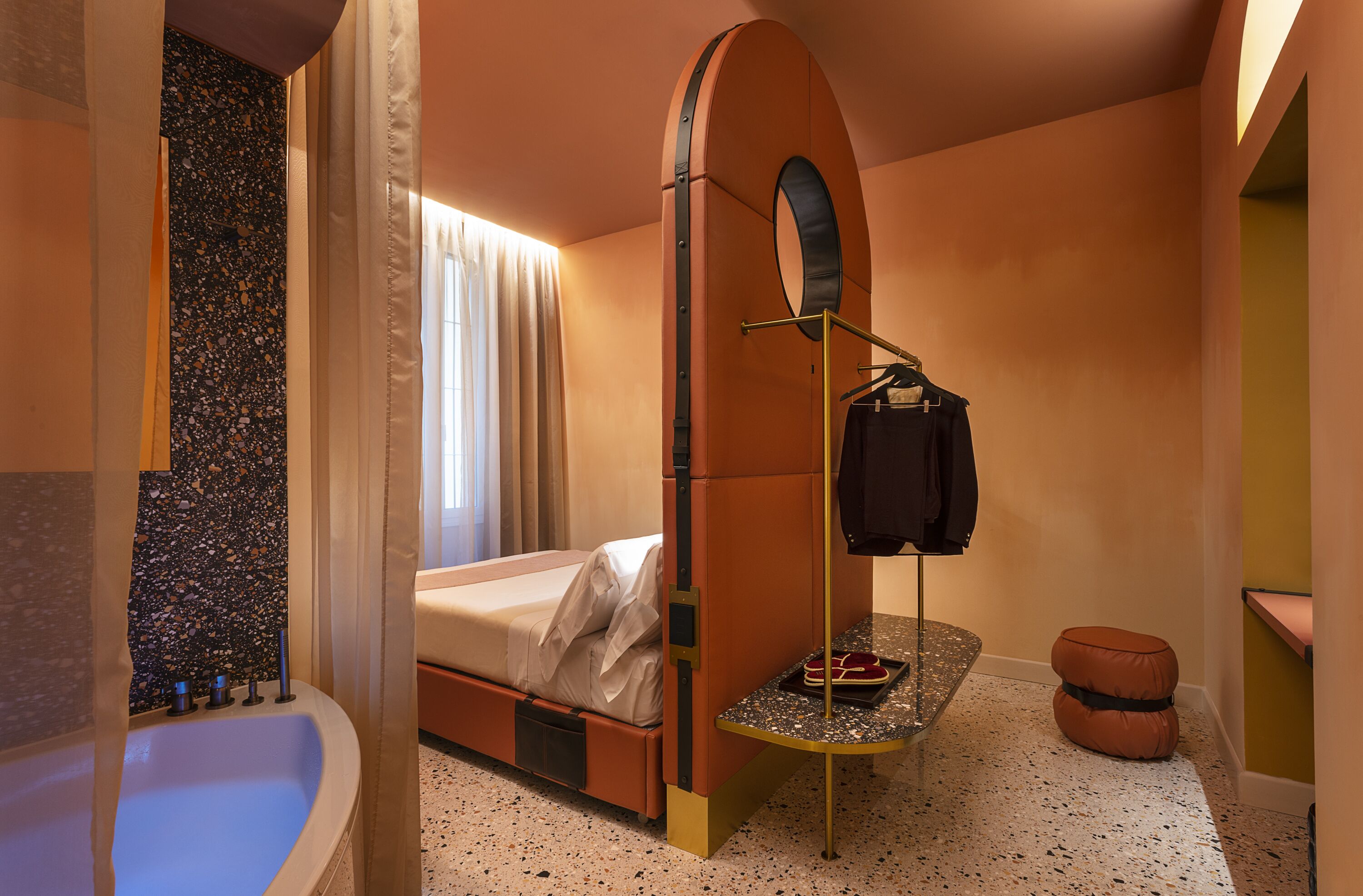 A compact hotel room with a bed, a small table with a tray, a hanging bag rack, a round mirror, and a small stool.