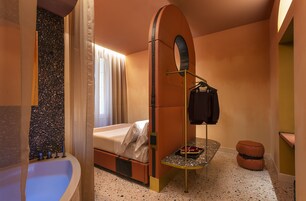 Een compacte hotelkamer met een bed, een klein tafeltje met dienblad, een kledingrek, een ronde spiegel en een klein krukje.