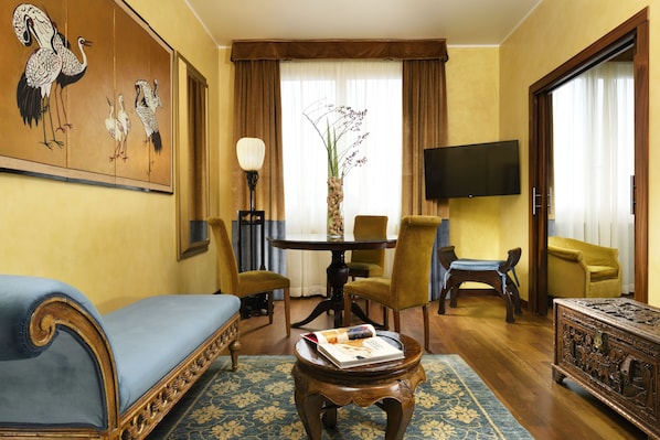 Un salon comprenant un canapé, des fauteuils, une table basse, un tableau, un téléviseur et une grande fenêtre avec des rideaux.