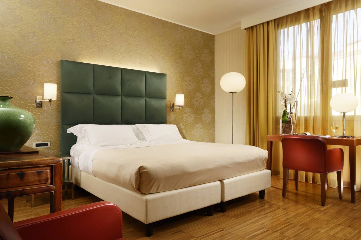 A hotel room with a large bed, a desk with a chair, a vase with flowers, and a lamp.