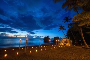 A beach at dusk with a row of candles on the sand, palm trees, and a small structure with lights. - Anse Chastanet Resort (Soufrière)