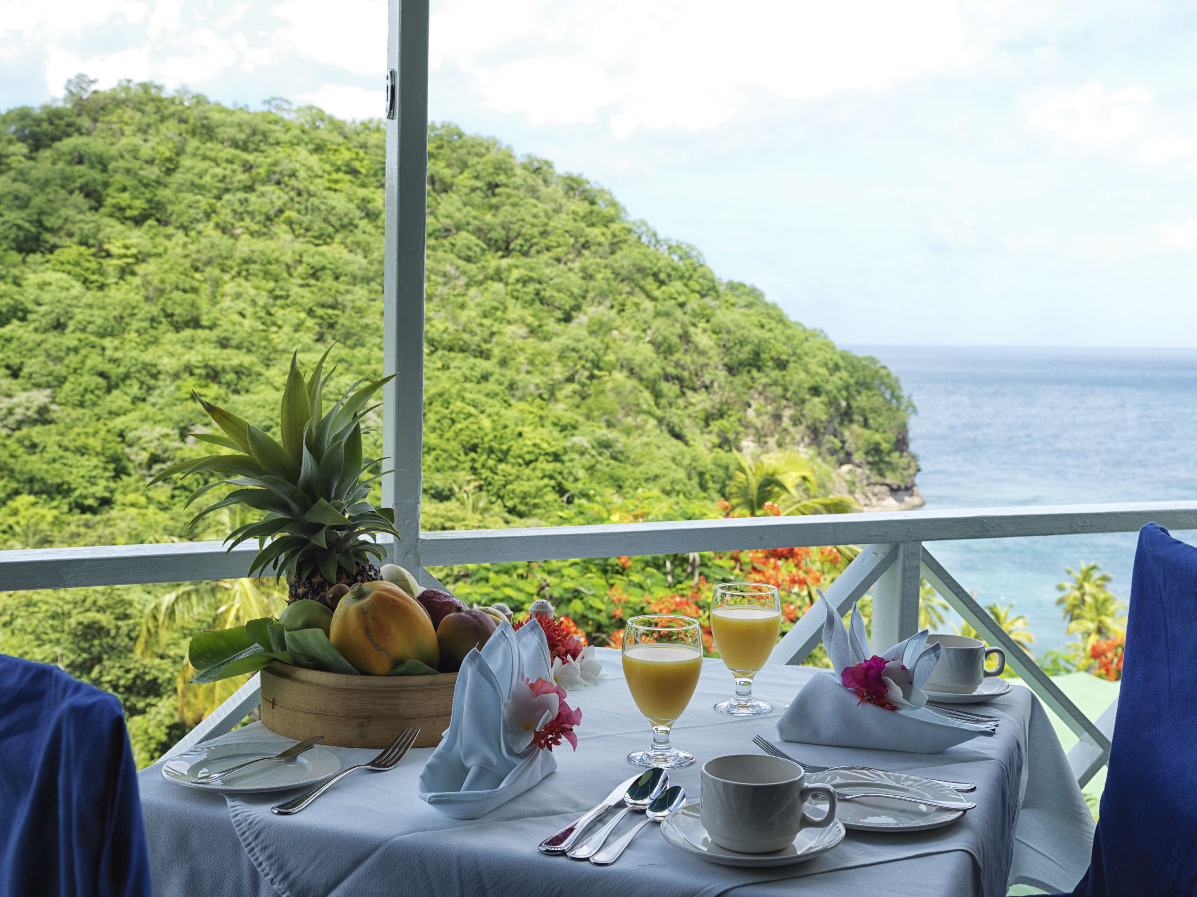 A table set with fruits, juice, and a cup, overlooking a lush green landscape and the ocean.