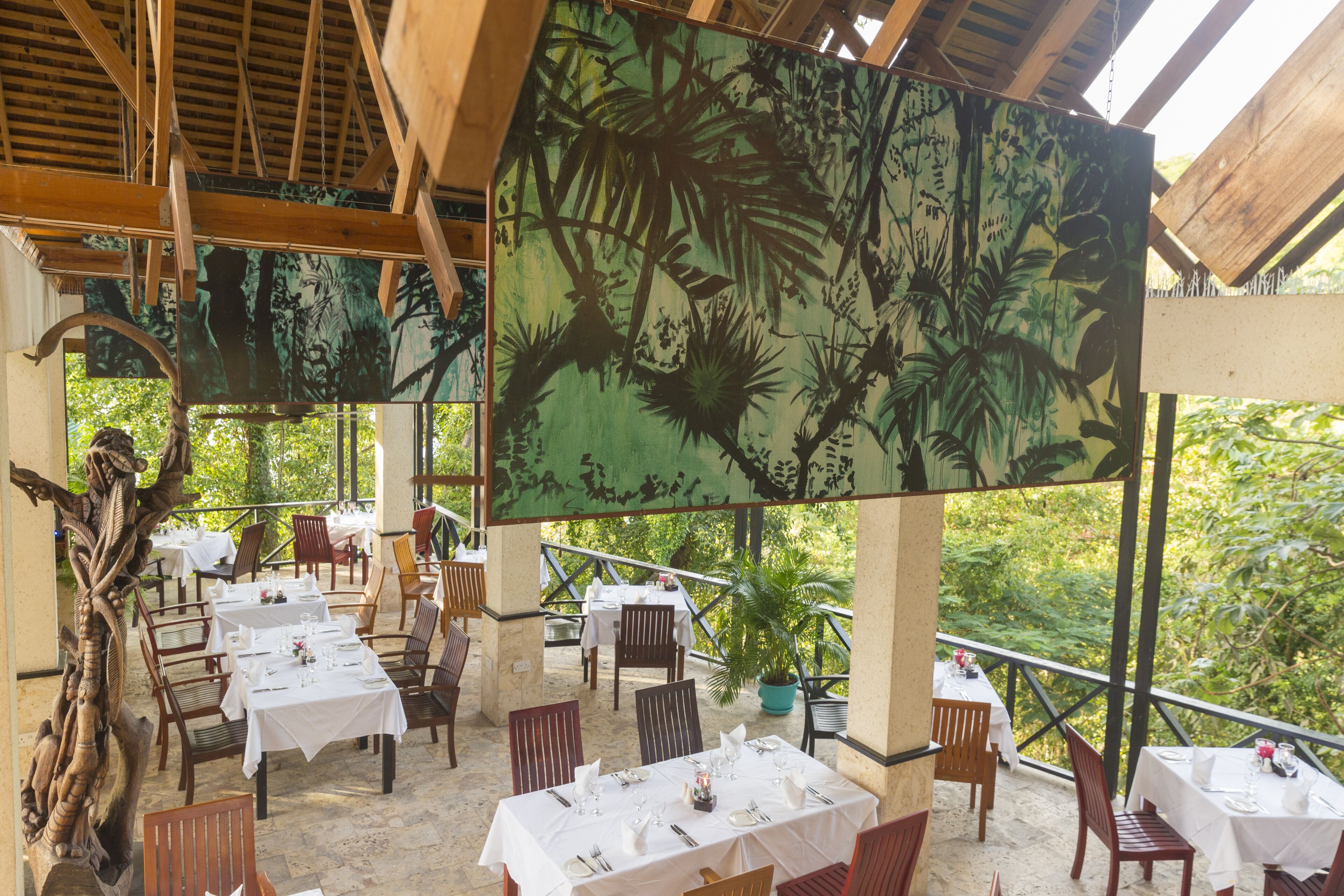 An outdoor dining area with tables set for two, surrounded by wooden chairs and a large tropical mural on the ceiling.