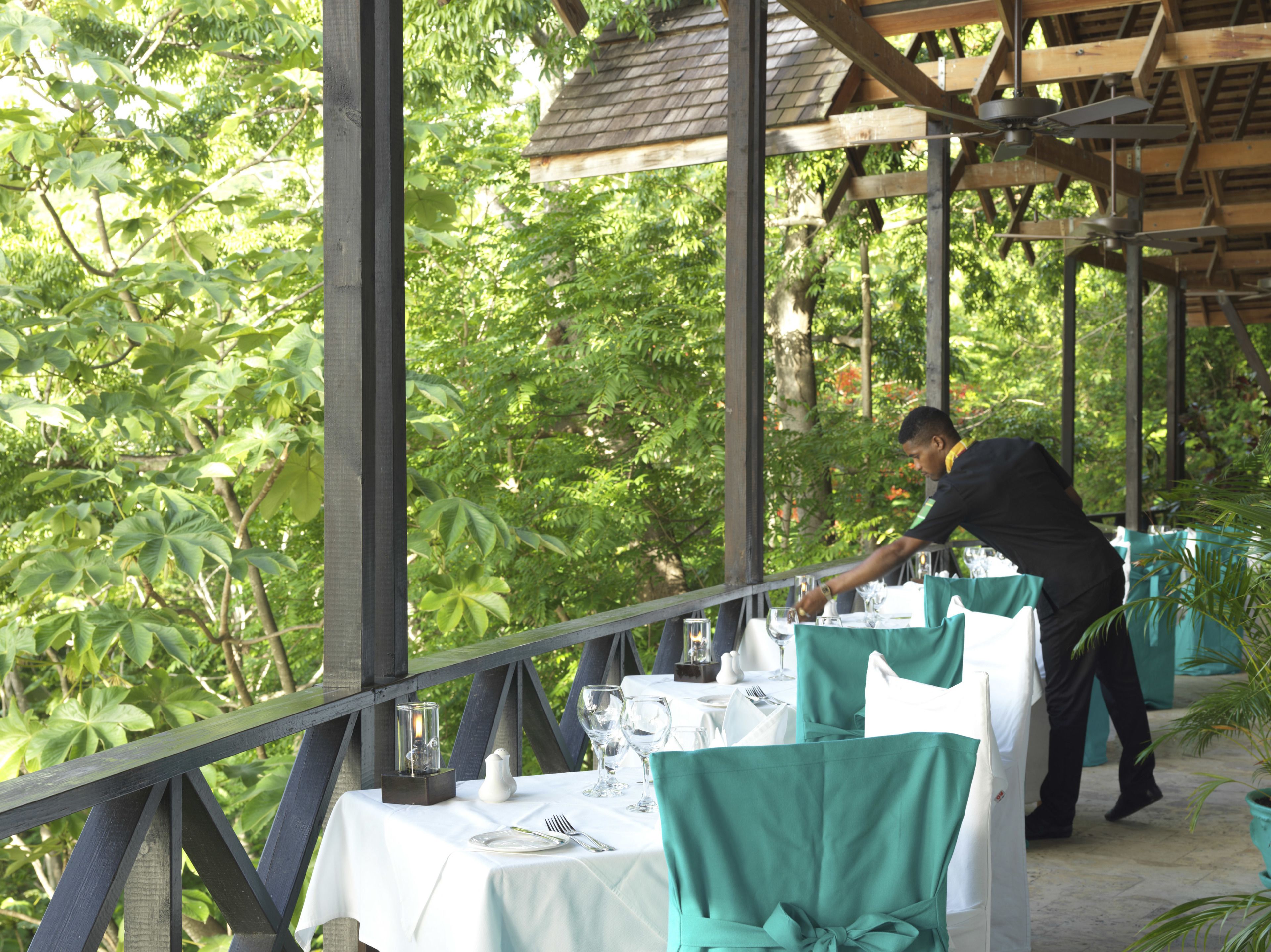 Un espacio de comedor al aire libre con mesas dispuestas para dos personas, rodeado de abundante vegetación.