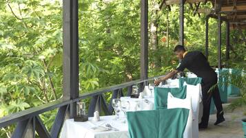 Un espacio de comedor al aire libre con mesas dispuestas para dos personas, rodeado de abundante vegetación.