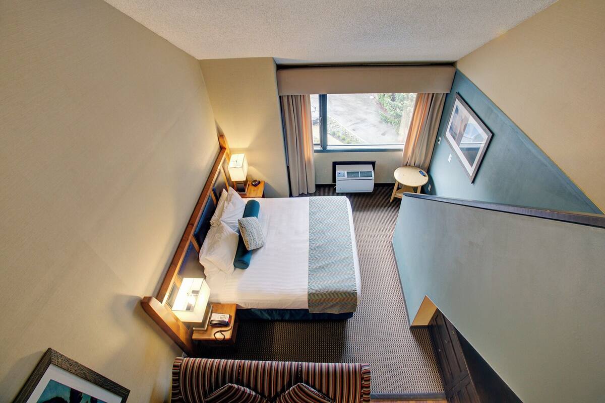 Habitación de hotel con cama, mesita de noche, lámpara, unidad de aire acondicionado y vistas al exterior.
