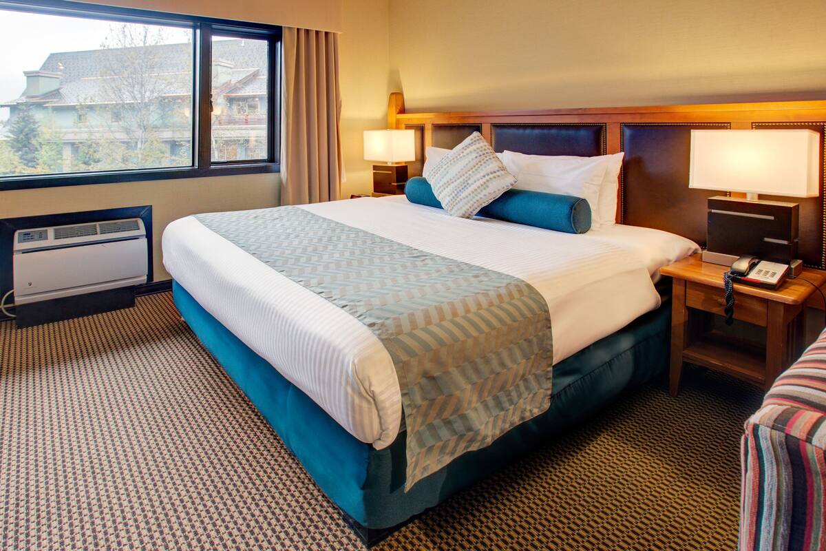 Habitación de hotel con una cama grande, una mesita de noche con lámpara, teléfono y ventana con cortinas.