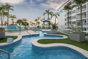Zona de piscina con sillas de descanso, palmeras y un edificio con balcones.