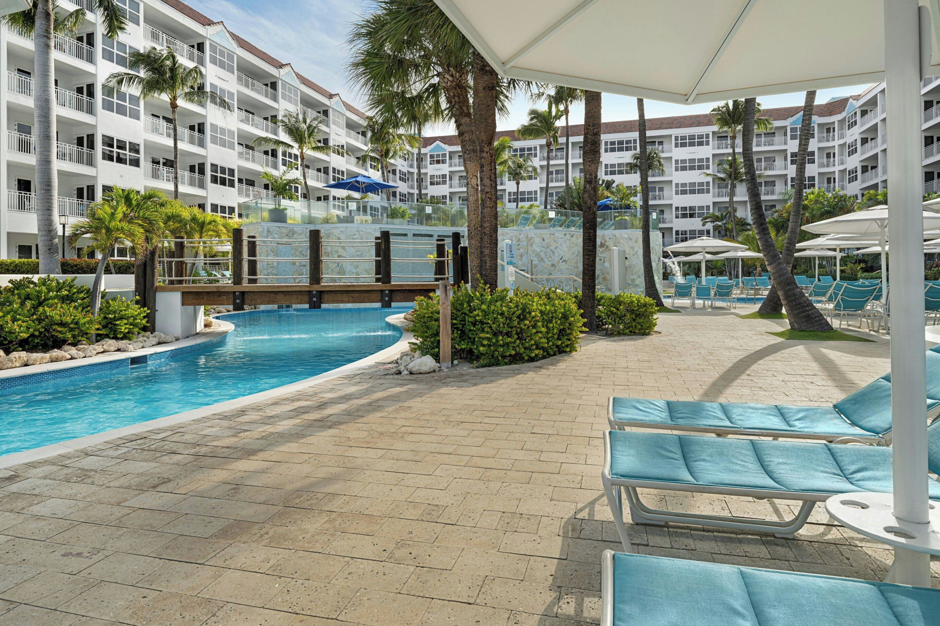 Un espace piscine d’hôtel avec des chaises longues, des palmiers et un immeuble à plusieurs étages en arrière-plan.