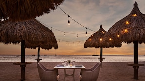 A beachside table with chairs, string lights, and thatched umbrellas.