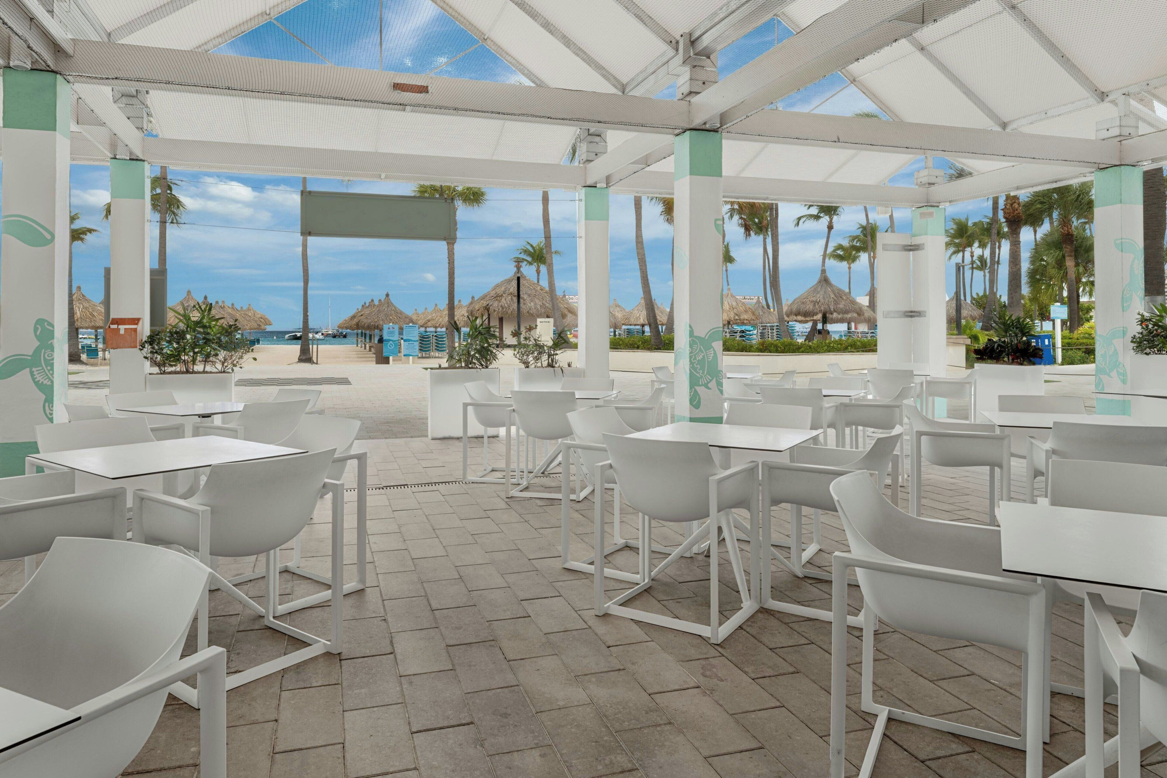 Un área de café al aire libre con mesas y sillas blancas, piso de baldosas y vista a la playa y palmeras.