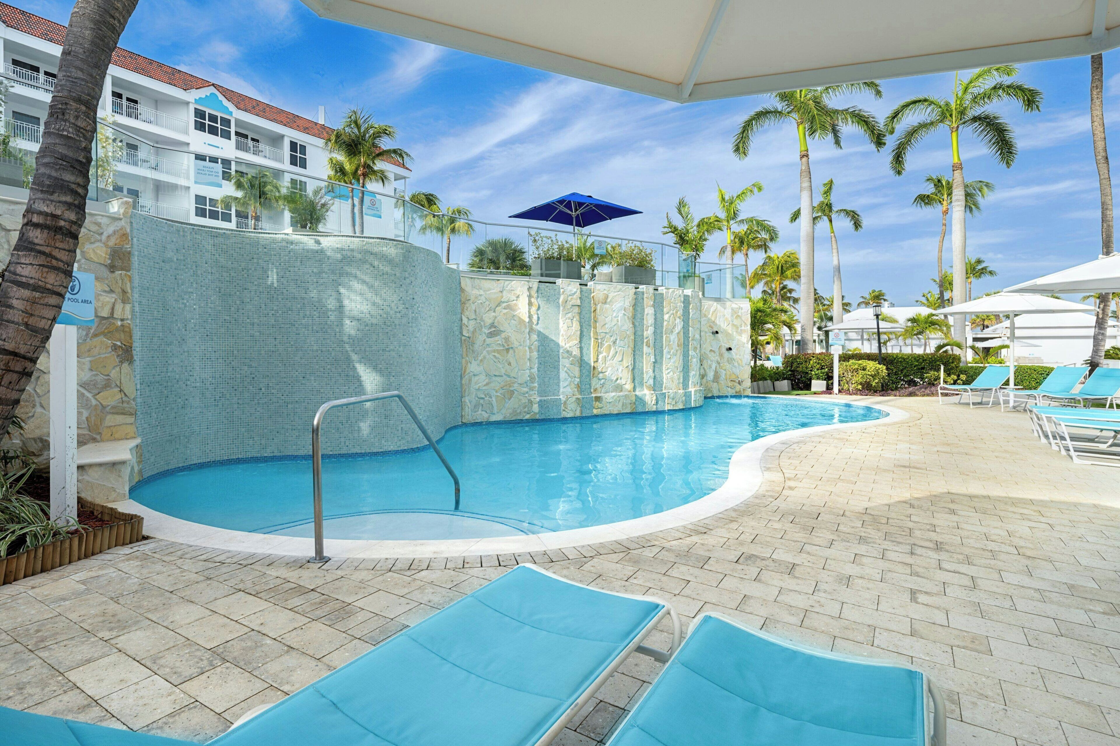 Un área de piscina con una piscina azul cristalina, sillones de descanso, palmeras y un muro de piedra.