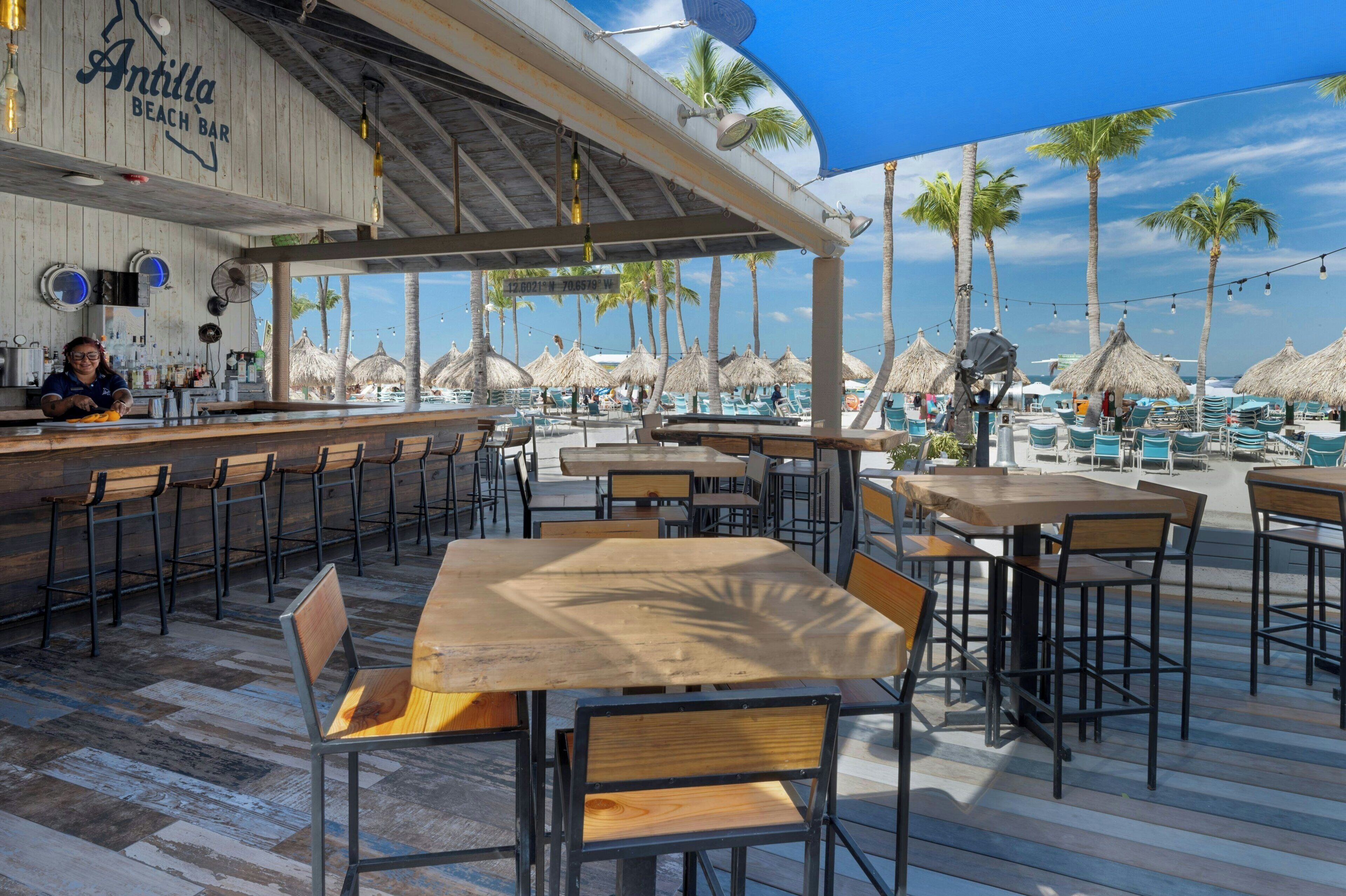 An outdoor bar area with wooden tables and chairs, a bartender, and thatched umbrellas.