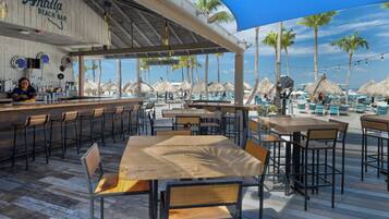 An outdoor bar area with wooden tables and chairs, a bartender, and thatched umbrellas.
